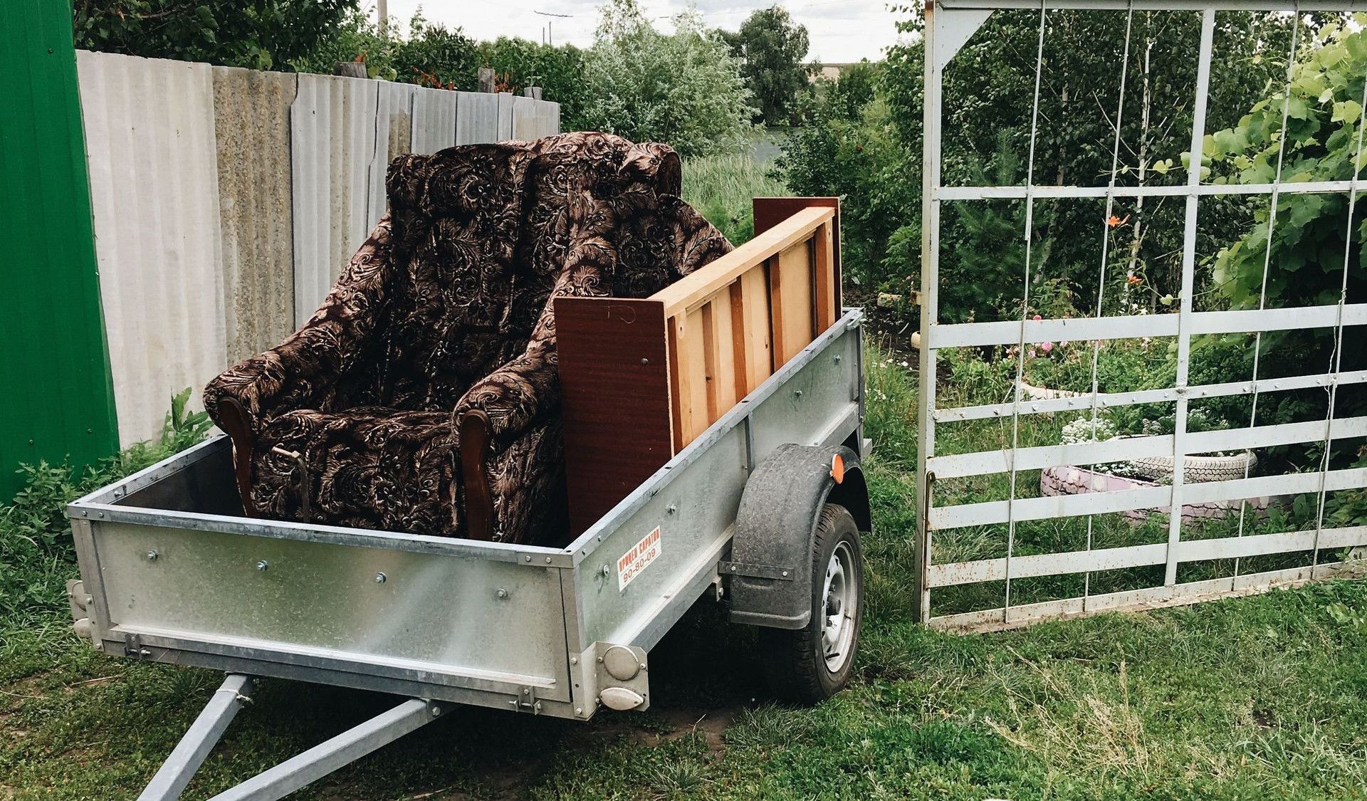 A rural setting with a trailer, vintage armchair, and a pug surrounded by greenery.