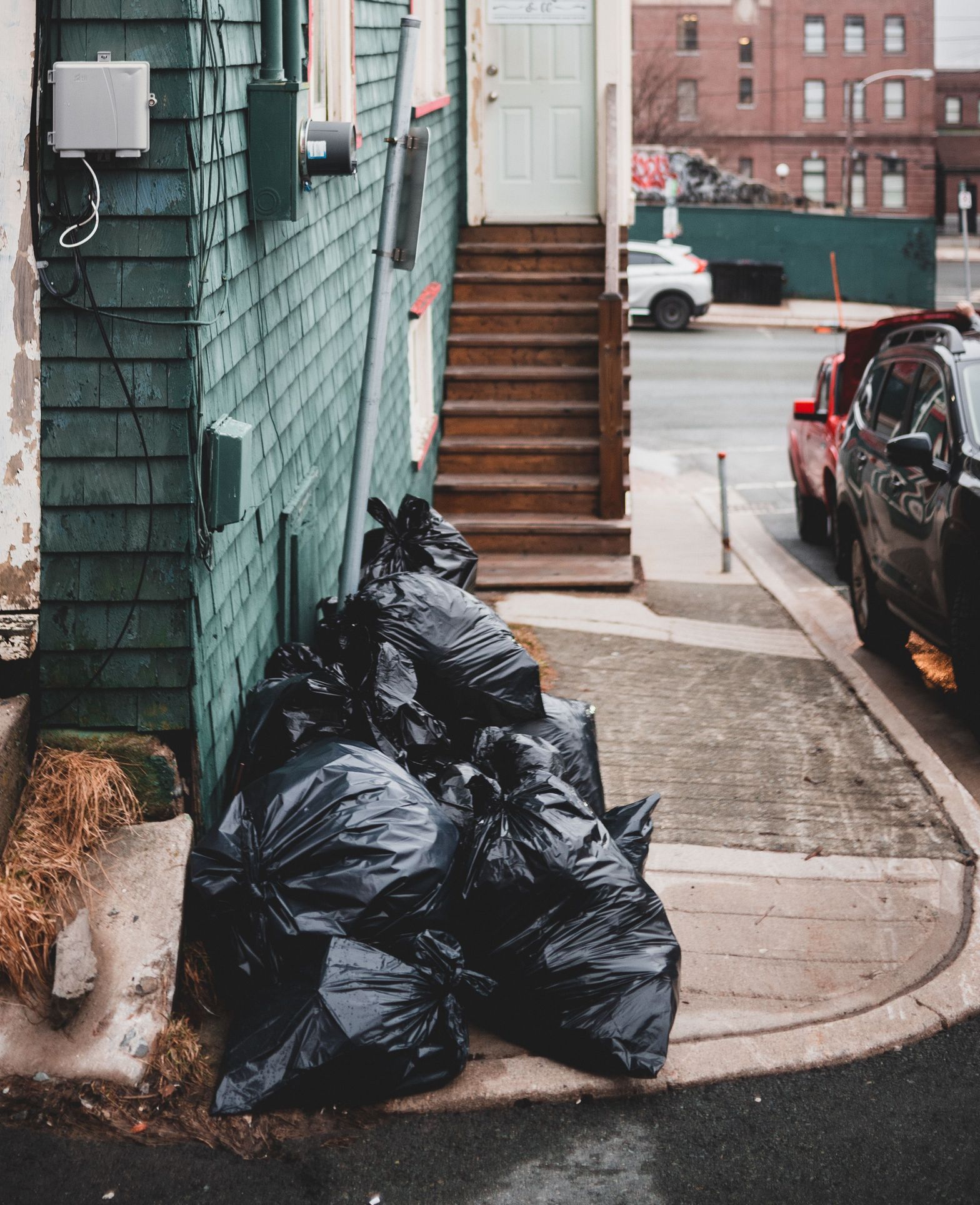 Black plastic garbage bags placed on sidewalk near colorful residential house with shabby stairs on street with buildings and cars